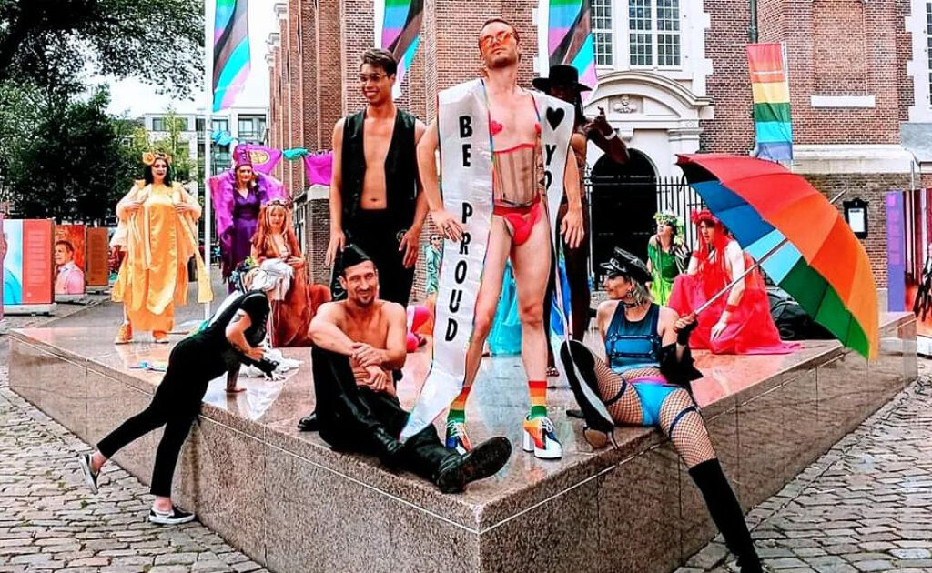 Photo of a group of drag performers with brightly coloured clothes and umbrellas sitting on a stone pedestal