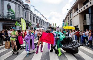 Photo of a group of drag activists on a public protests, with a bright red flag with the text "House of Extinction"
